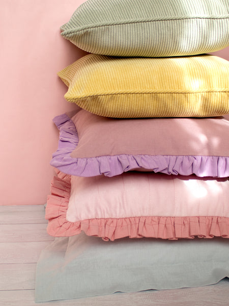 Five different colourful cushions are placed on top of each other on a white wooden floor and a pink wall.