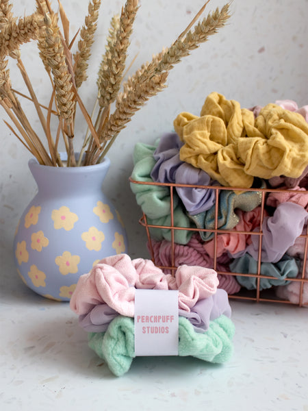 A bundle of three scrunchies wrapped together with a 'Peachpuff Studios" label. A basket of scrunchies in the background with a handpainted vase that contains dried wheat.