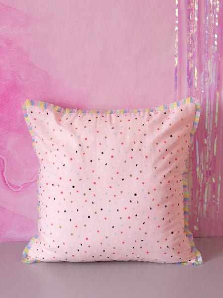 A pink cushion with pastel rainbow fringe in front of a marbled pink wall. 