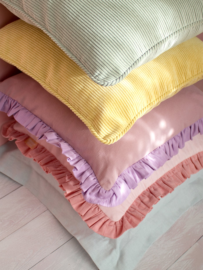 Five different colourful cushions are placed on top of each other on a white wooden floor and a pink wall.