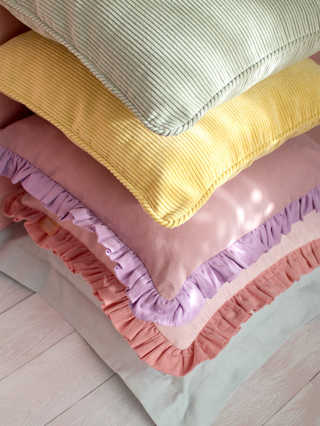 Five different colourful cushions are placed on top of each other on a white wooden floor and a pink wall.