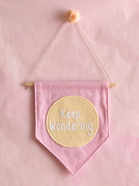 A pink canvas banner that reads 'Keep Wondering' in a yellow circle is hung on a pink wall.