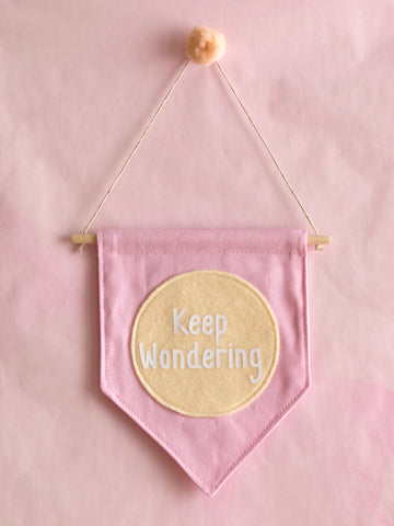 A pink canvas banner that reads 'Keep Wondering' in a yellow circle is hung on a pink wall.