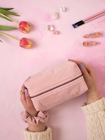 A pair of female hands holding a pink corduroy zip pouch on a pink-marbled floor. Flowers, crystals, makeup brushes and hair clips offset at the top.