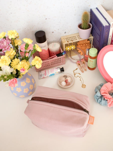 A pink corduroy zip pouch opened on a busy dressing table filled with beauty products, books and a vase of flowers.