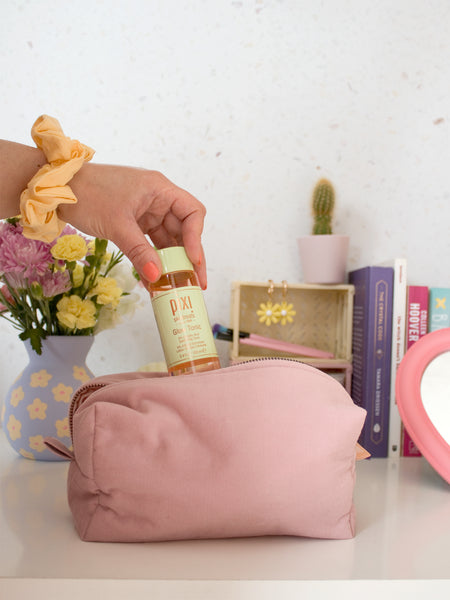A female hand with a scrunchie on her wrist puts a bottle into a pink corduroy makeup bag.
