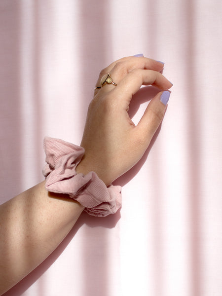 A pink corduroy scrunchie on a female wrist, who is wearing a ring and purple nail varnish.