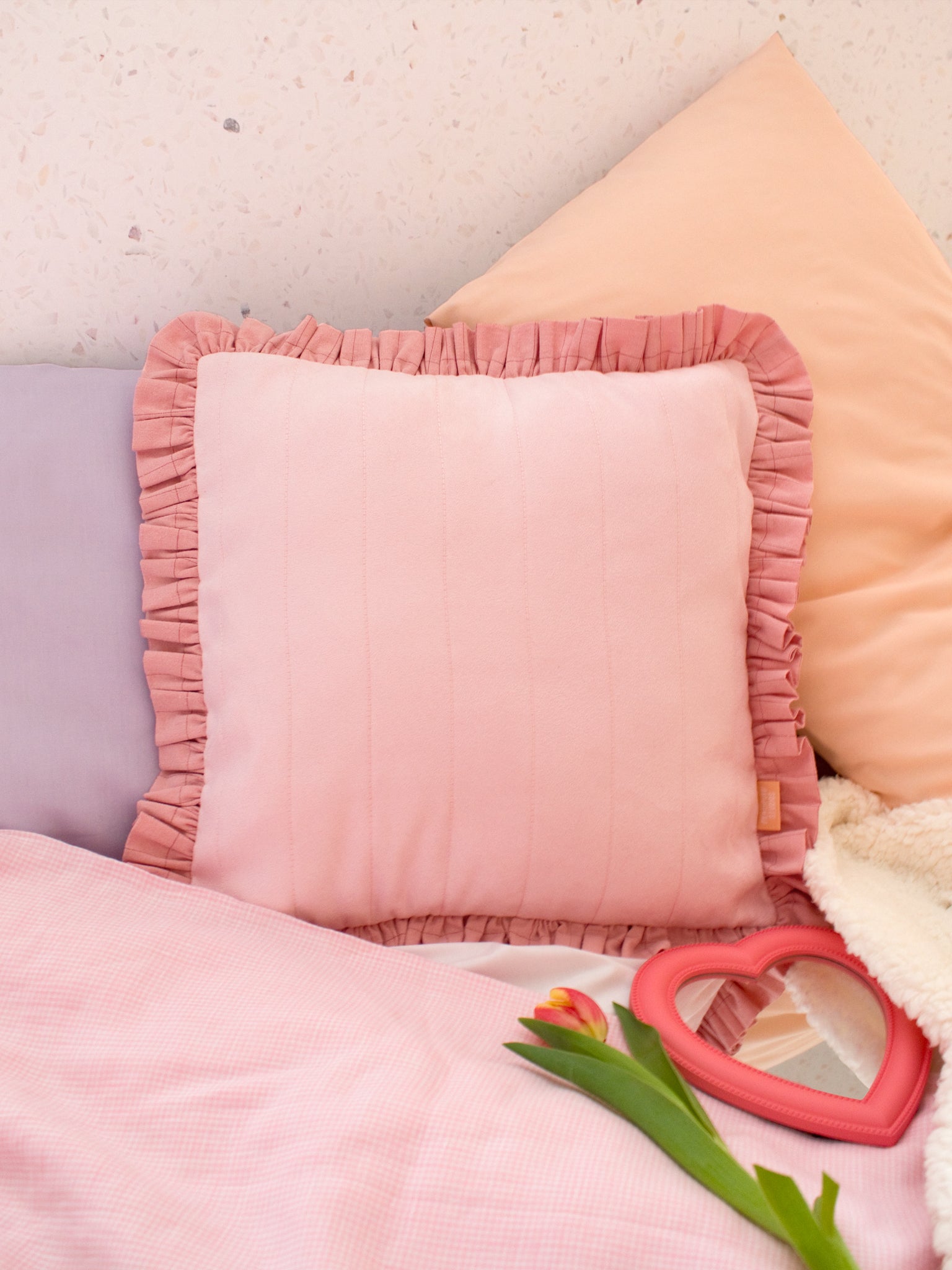 A pink faux suede ruffled cushion styled on a bed with pastel pillowcases and gingham sheets.