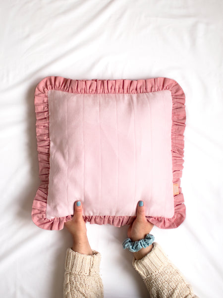 A pink faux suede ruffle cushion on the centre of a white bedsheet held at the bottom by a pair of female hands.