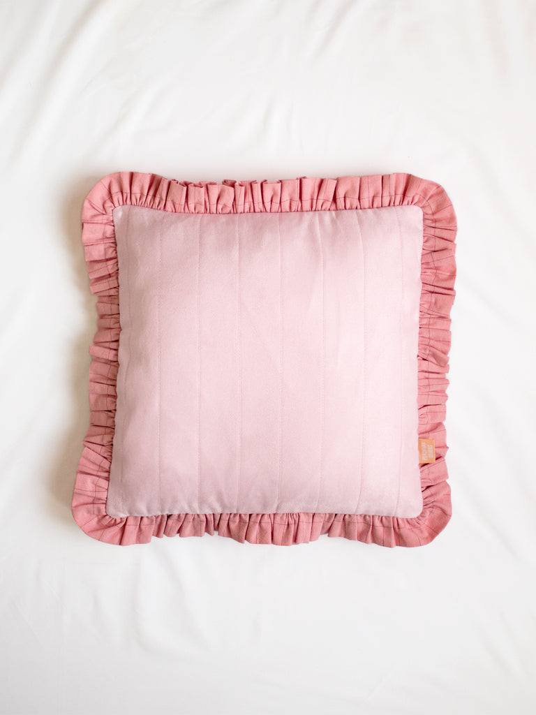 A pink faux suede ruffle cushion on the centre of a white bedsheet.