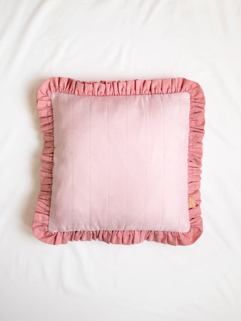 A pink faux suede ruffle cushion on the centre of a white bedsheet.