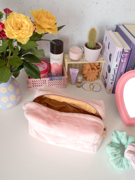 A pink plush zip pouch opened on a busy dressing table filled with beauty products, books and a vase of flowers.