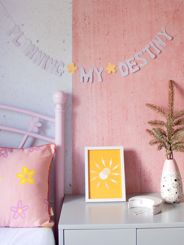 A bedroom with a pink flower bed and grey bedside table displaying a purple texted garland above that reads 'Planning My Destiny'.