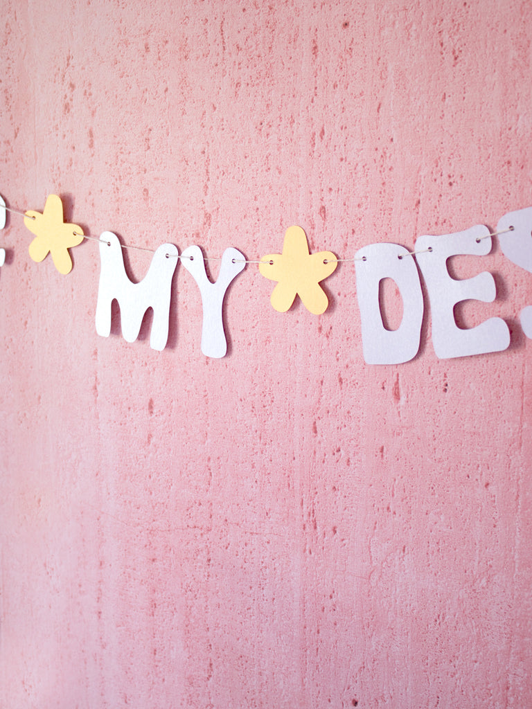 A paper garland that's made of purple text and yellow flowers hung up on a pink-textured wall.