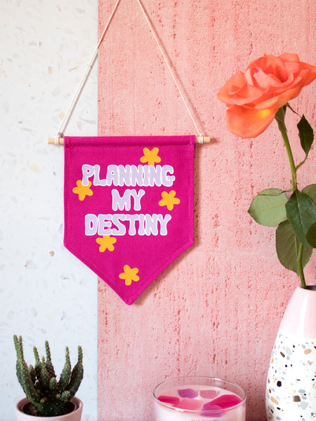 A pink canvas banner that reads Planning My Destiny with purple lettering is hung on a white and pink textured wall surrounded by a cactus, flowers and a candle.