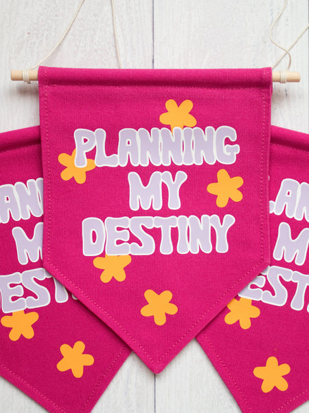 Three pink canvas banners that read Planning My Destiny with purple lettering are laid on top of each other on a white wooden floor.