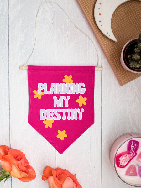 A pink canvas banner that reads Planning My Destiny with purple lettering is placed on a white wooden floor with flowers, a candle and trinkets surrounding it.