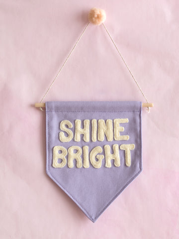 A purple canvas banner that reads Shine Bright in yellow fleece lettering is hung on a pink wall.