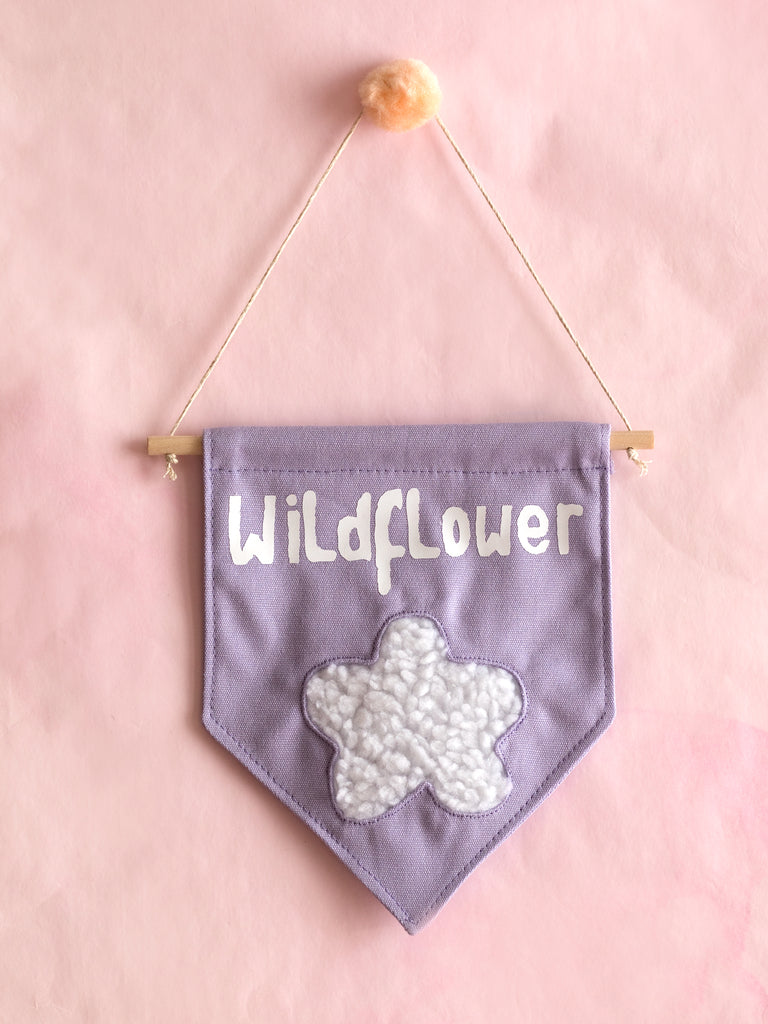 A purple canvas banner that reads Wildflower with a fluffy white flower at the bottom is hung on a pink wall.
