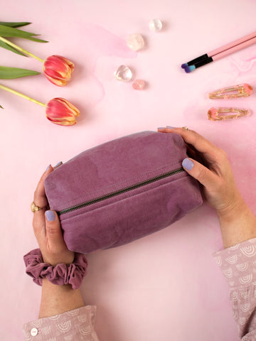 A pair of female hands holding a purple corduroy zip pouch on a pink-marbled floor. Flowers, crystals, makeup brushes and hair clips offset at the top.