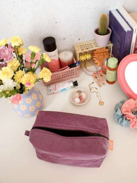 A purple corduroy zip pouch opened on a busy dressing table filled with beauty products, books and a vase of flowers.