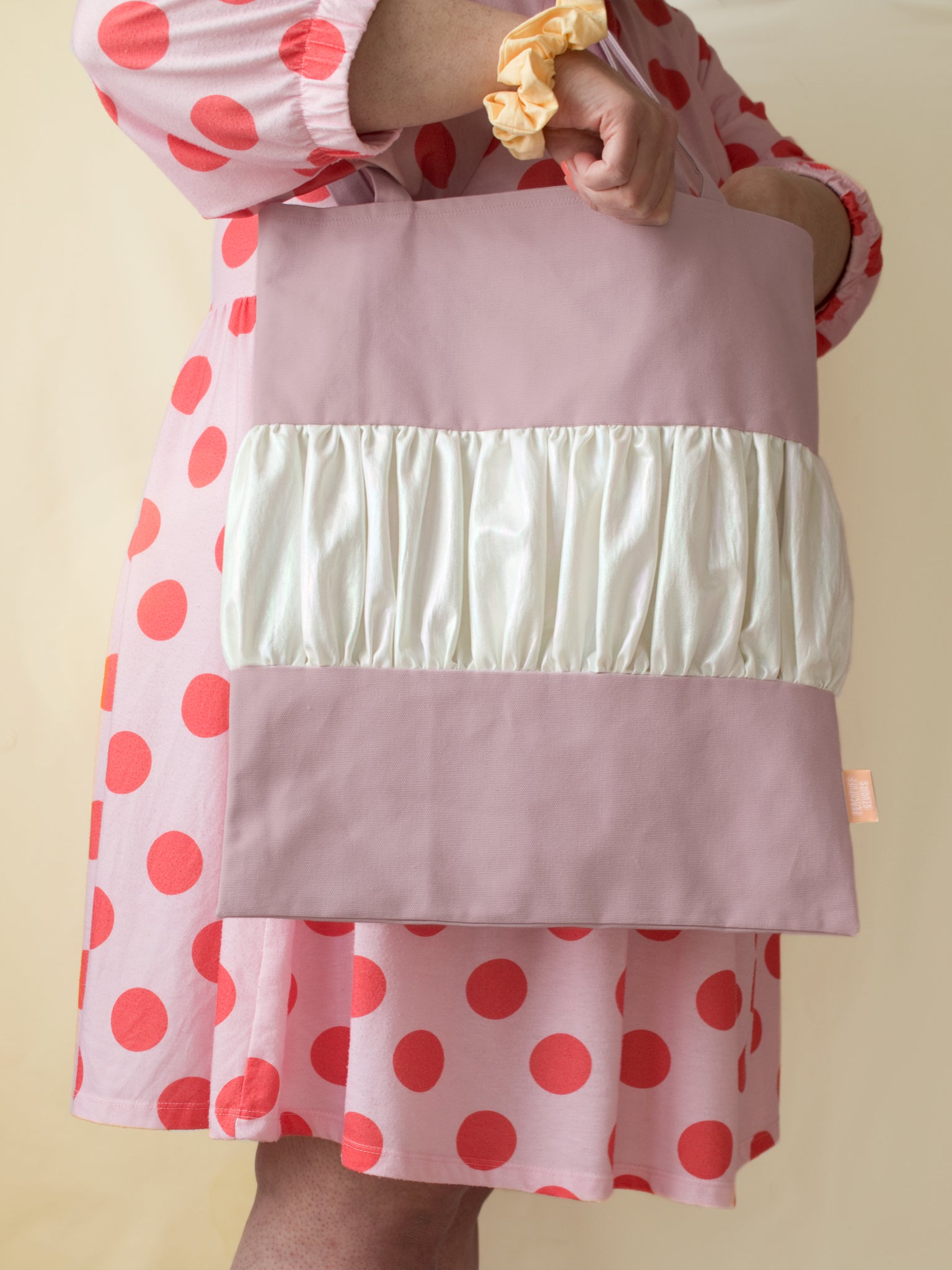 A purple tote bag with a centre ruffle is worn on the shoulder of a female who is wearing a pink and red polka dot dress.