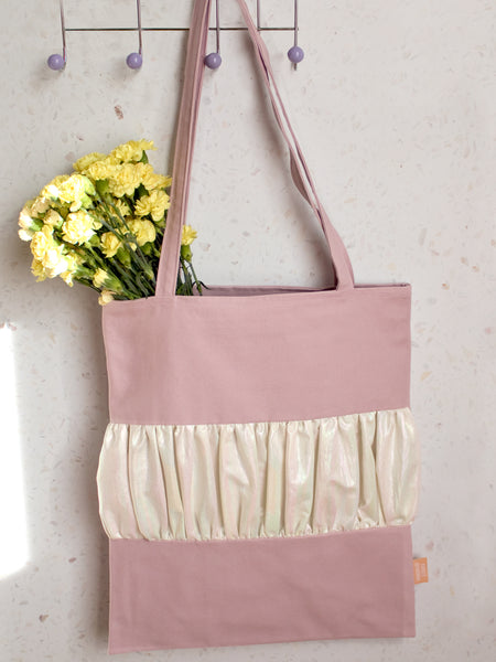A purple tote bag with a centre ruffle holding yellow flowers is hung on a peg behind a terrazzo wall.