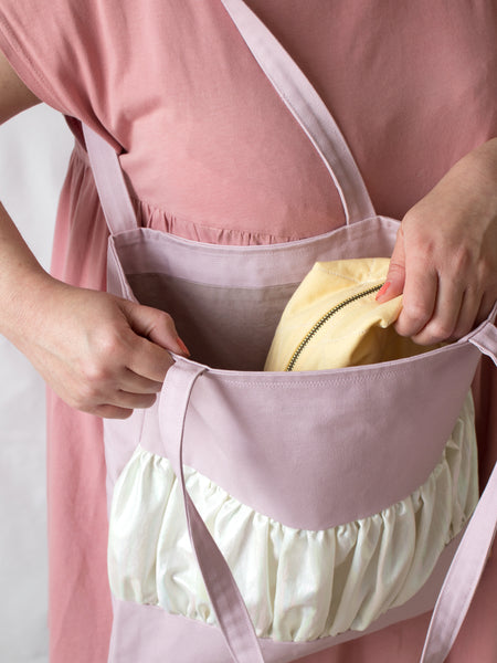 A purple tote bag with a centre ruffle is worn on the shoulder of a female who is wearing a pink dress. She is putting a yellow makeup bag into the tote.