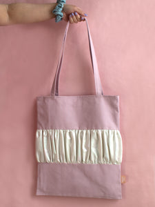 A female arm holds a purple tote bag with a ruffle in the middle against a pink foreground.