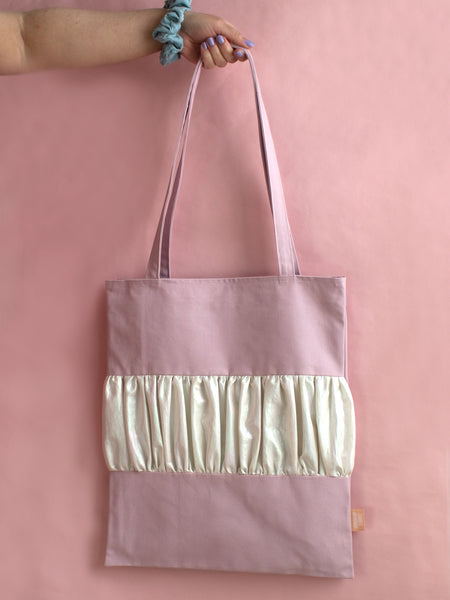 A female arm holds a purple tote bag with a ruffle in the middle against a pink foreground.