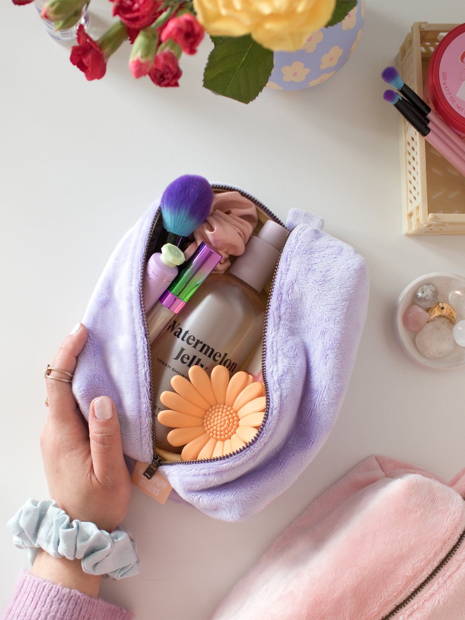 A female hand with a scrunchie on her wrist opens a purple plush makeup bag to reveal beauty products.