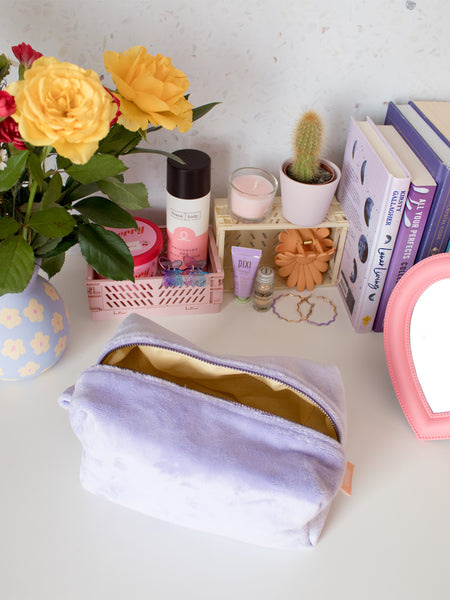 A purple plush zip pouch opened on a busy dressing table filled with beauty products, books and a vase of flowers.