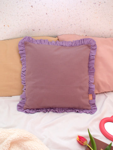 A purple ruffle cushion styled on a bed with pastel pillowcases and fluffy sheets.