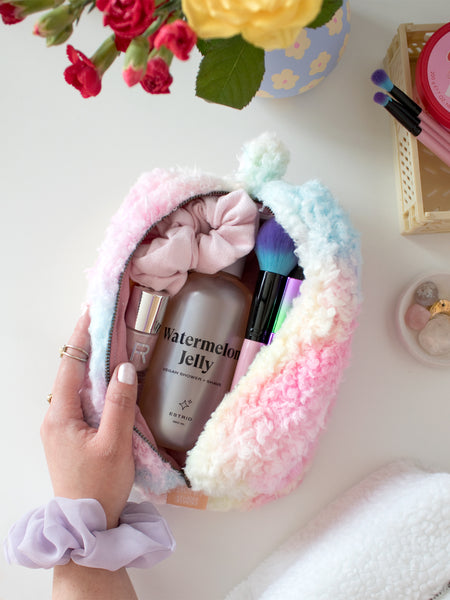 A female hand with a scrunchie on her wrist opens a rainbow fluffy makeup bag to reveal beauty products.