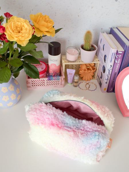 A rainbow fluffy zip pouch opened on a busy dressing table filled with beauty products, books and a vase of flowers.