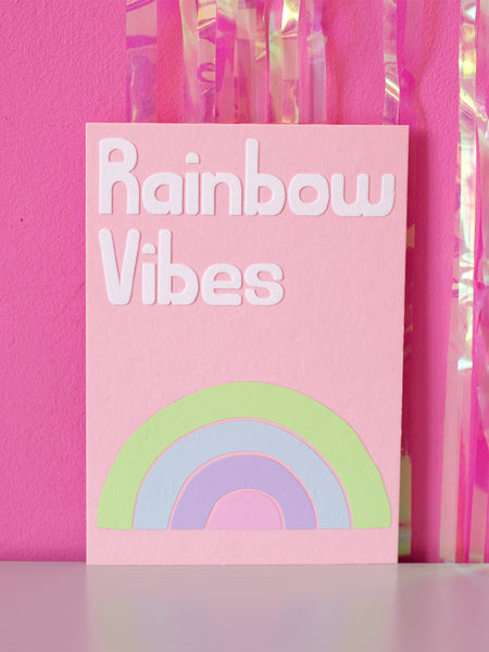 Art print with Rainbow Vibes in flocked font written on top of a drawn rainbow made up of four stripes of colour, sitting in front of a pink wall.
