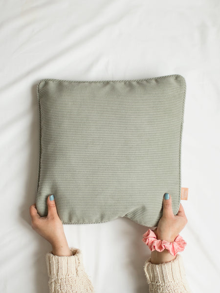 A sage green corduroy cushion on the centre of a white bedsheet held at the bottom by a pair of hands.