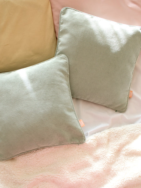 Two sage green corduroy cushions styled on a bed with pastel pillowcases and fluffy sheets.