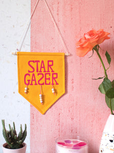 A yellow canvas banner that reads Star Gazer and is decorated with hanging beads is hung on a white and pink textured wall surrounded by a cactus, flowers and a candle.