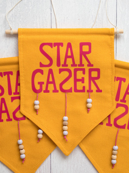 Three yellow canvas banner that reads Star Gazer and is decorated with hanging beads are laid on top of each other on a white wooden floor.