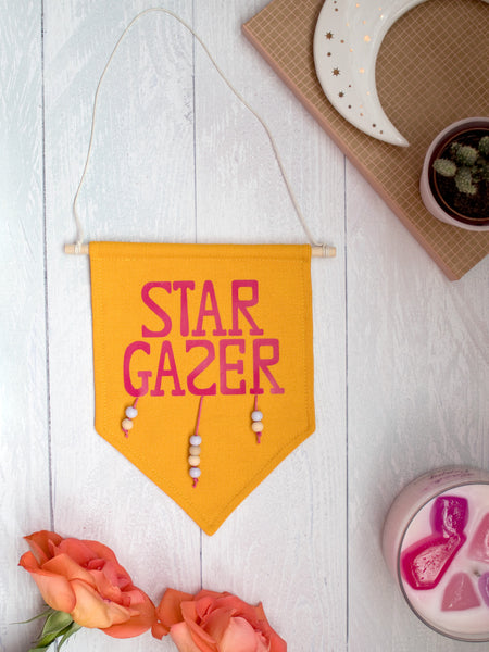 A yellow canvas banner that reads Star Gazer and is decorated with hanging beads is placed on a white wooden floor with flowers, a candle and trinkets surrounding it.