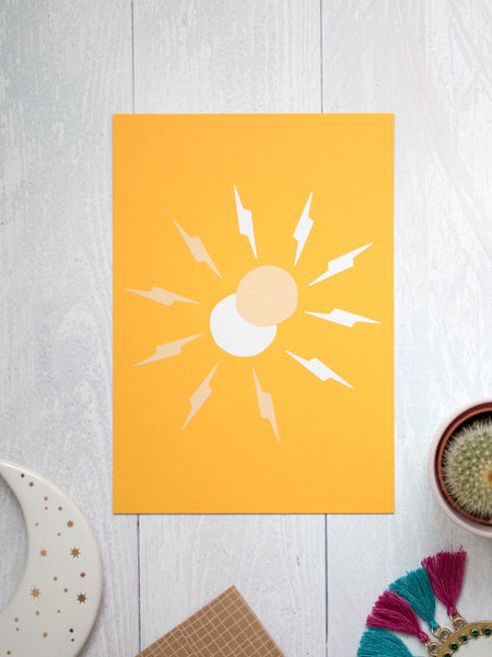 An art print of a simple beige and white sun with thunderbolt rays overlapping lay on a white wooden floor. Trinkets and a cactus lay offset at the bottom.