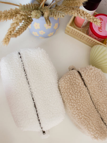 Two teddy makeup bags, one white and one cream, are on a table from a top view. There are also dried flowers in a blue-painted pot, a shell candle and beauty products in a small crate.