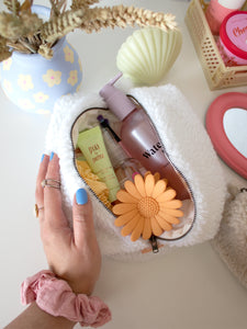 A female hand with a scrunchie on her wrist opens a white, fluffy makeup bag to reveal beauty products.
