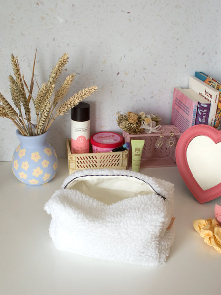 A white, fluffy zip pouch opened on a busy dressing table filled with beauty products, books and a vase of flowers.