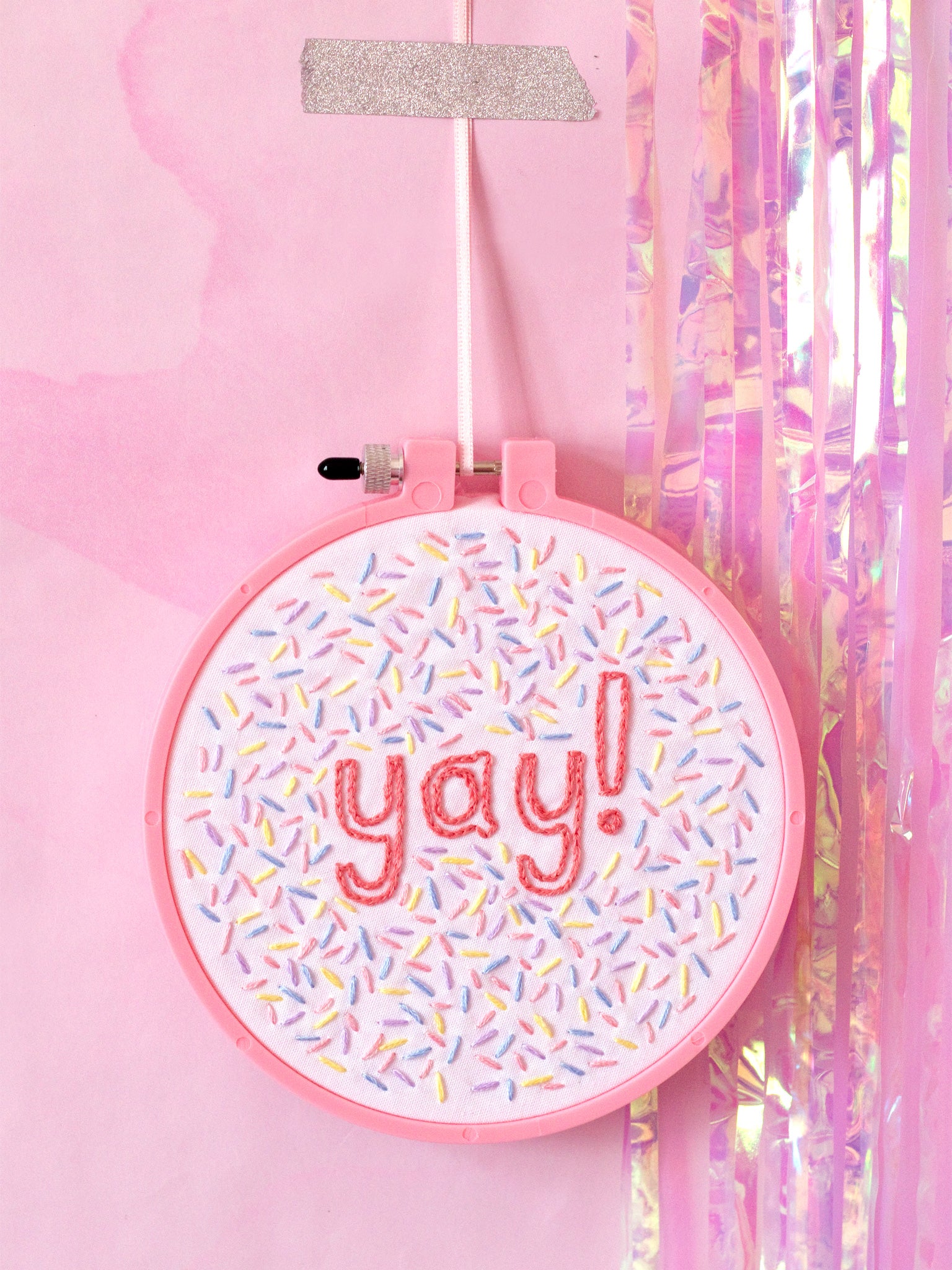 A pink embroidery hoop with white fabric stitched with yay and colourful sprinkles. Hung on a pink marbled wall.