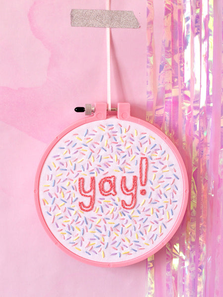 A pink embroidery hoop with white fabric stitched with yay and colourful sprinkles. Hung on a pink marbled wall.