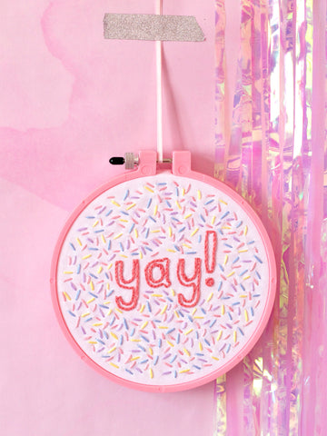 A pink embroidery hoop with white fabric stitched with yay and colourful sprinkles. Hung on a pink marbled wall.