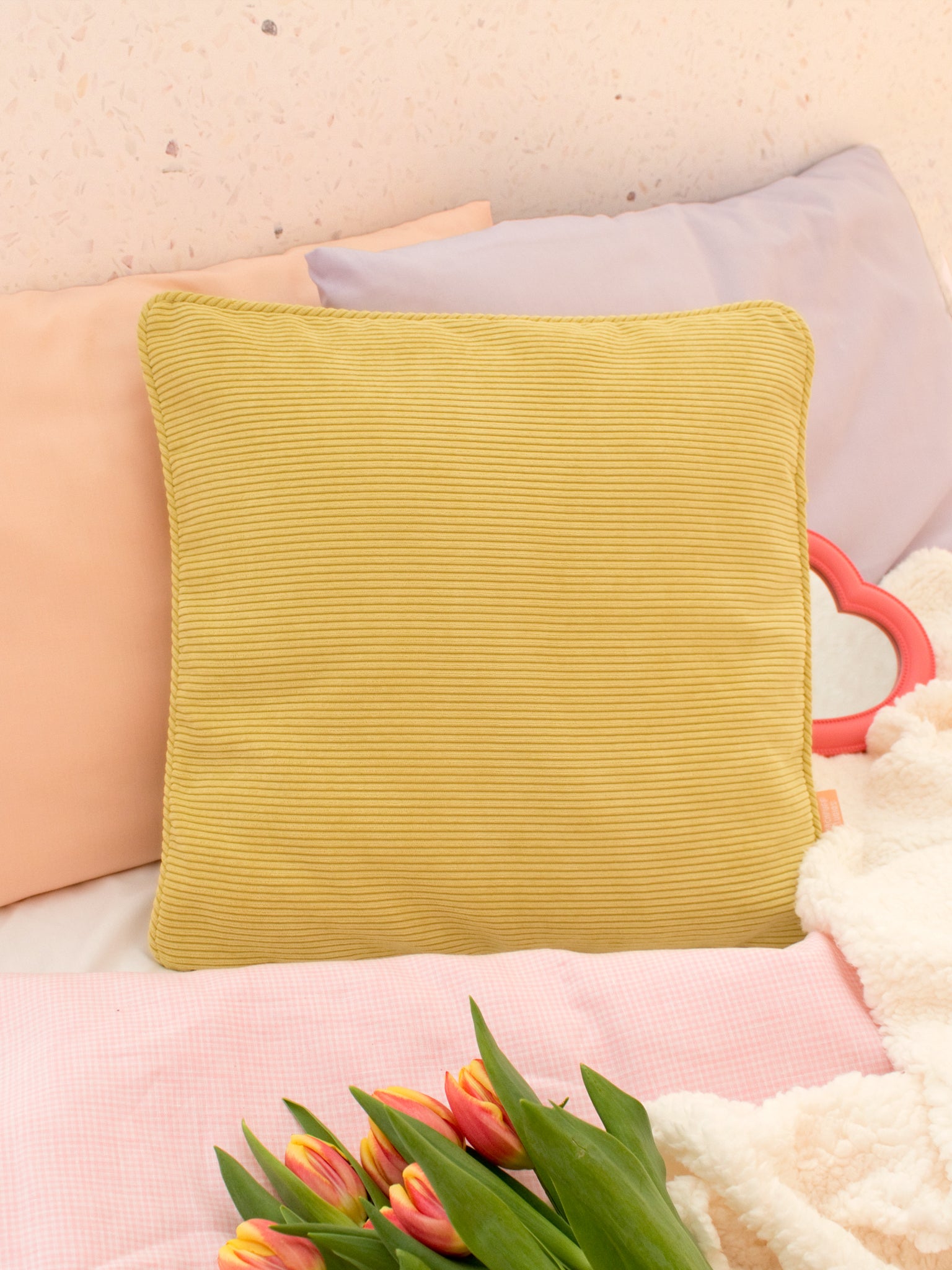 A yellow corduroy cushion styled on a bed with pastel pillowcases and gingham sheets.