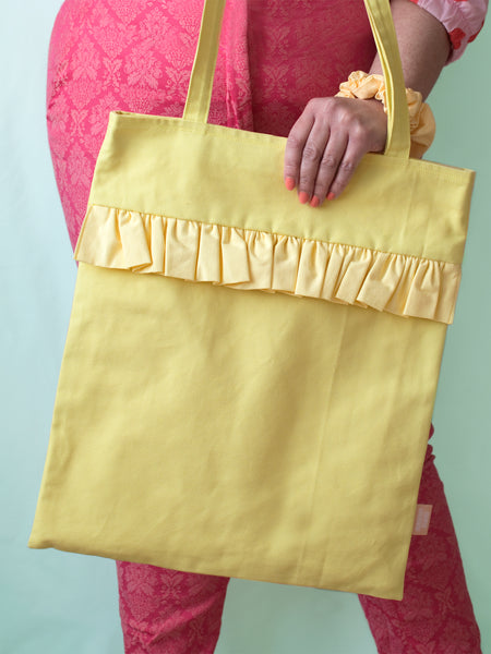A yellow ruffle tote bag is held against a pair of pink patterned trousers.